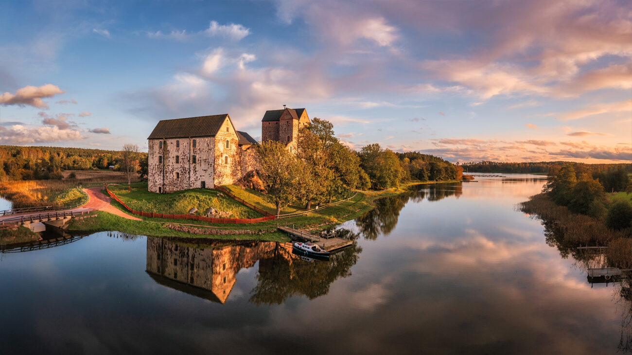 zwei größere, helle Gebäude auf einer grünen Hügelhalbinsel am Wasser mit rosa Wolken im Hintergrund