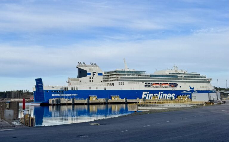 Large white and blue First Lines ferry docked at a harbor with calm water reflecting the ship, under a partly cloudy sky.