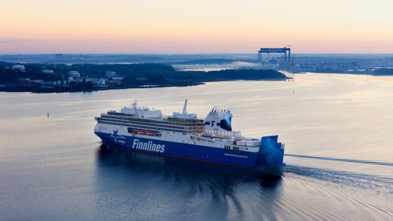 Large Finnlines cargo ship sailing on calm water during sunset with industrial buildings and a large crane visible in the distant background.