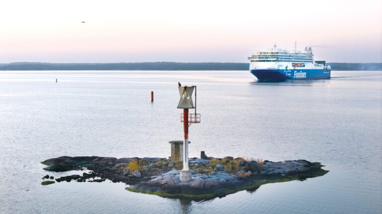 A small rocky islet with a metal navigation beacon stands in calm, pastel-colored dawn waters while a large blue-and-white passenger ferry approaches in the distance and channel buoys mark the route.