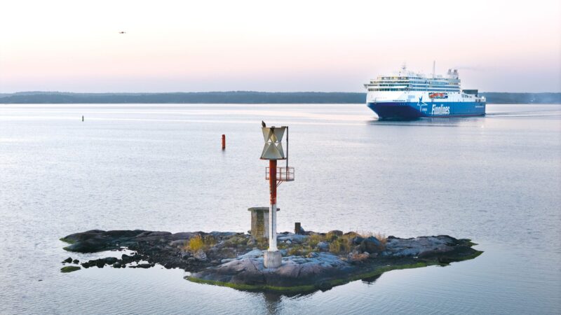 A small rocky islet with a metal navigation beacon stands in calm, pastel-colored dawn waters while a large blue-and-white passenger ferry approaches in the distance and channel buoys mark the route.
