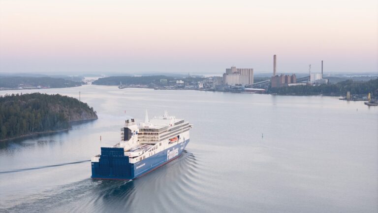 Aerial view of a large blue-and-white passenger ferry moving through calm coastal waters at dawn, leaving a V-shaped wake as it passes wooded islands toward an industrial shoreline with power-plant buildings under a pale pink and blue sky.
