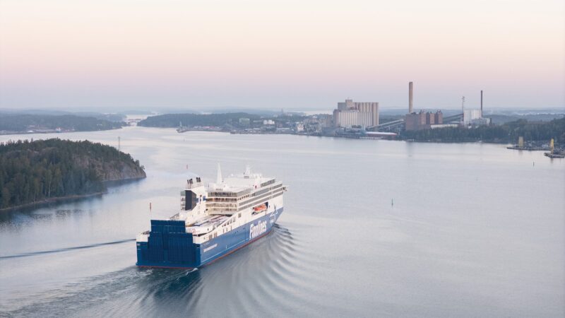 Aerial view of a large blue-and-white passenger ferry moving through calm coastal waters at dawn, leaving a V-shaped wake as it passes wooded islands toward an industrial shoreline with power-plant buildings under a pale pink and blue sky.