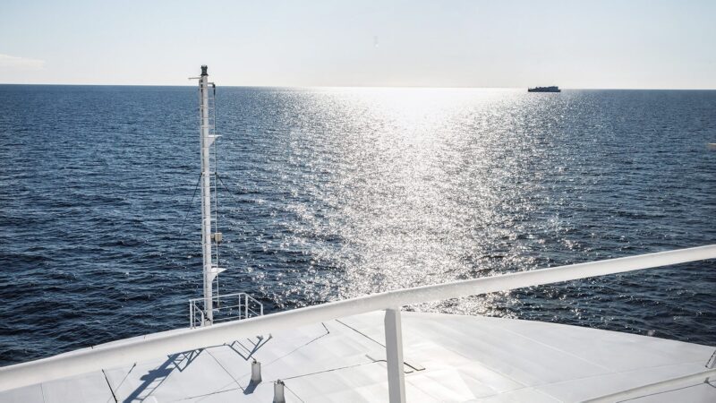View from a ship's bow over a calm, sunlit sea with the ship's white deck and railing in the foreground, a tall mast to the left and a small vessel on the distant horizon reflecting sunlight