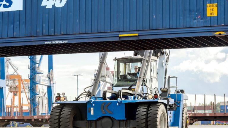 Front view of a large blue container handler (reach stacker) with heavy tires and a lifting spreader holding a shipping container at a busy port, with gantry cranes and stacked containers in the background under a cloudy sky.