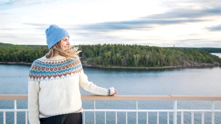 Person in a white patterned Nordic sweater and blue knit hat stands on a ferry deck, resting a hand on the railing and gazing toward a tree‑covered island across calm water under a pale, cloud‑streaked sky.