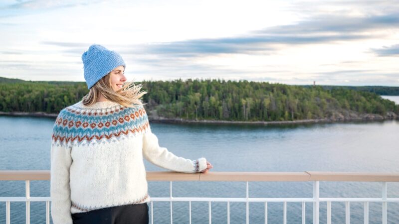 Person in a white patterned Nordic sweater and blue knit hat stands on a ferry deck, resting a hand on the railing and gazing toward a tree‑covered island across calm water under a pale, cloud‑streaked sky.