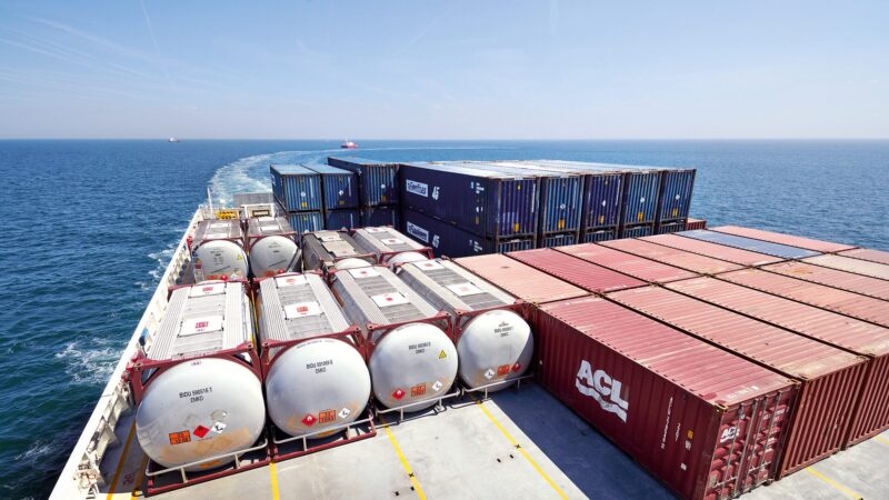 View from the deck of a container ship carrying rows of cylindrical tank containers and stacked rectangular shipping containers under a clear blue sky, with the ship's wake trailing across the calm sea.