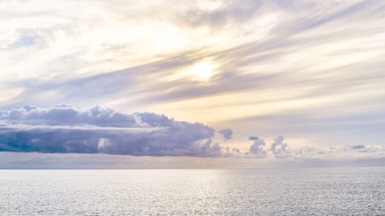 Calm sea under a pastel sky at sunrise, the sun partially hidden by wispy and darker clouds with soft light reflecting on the water.