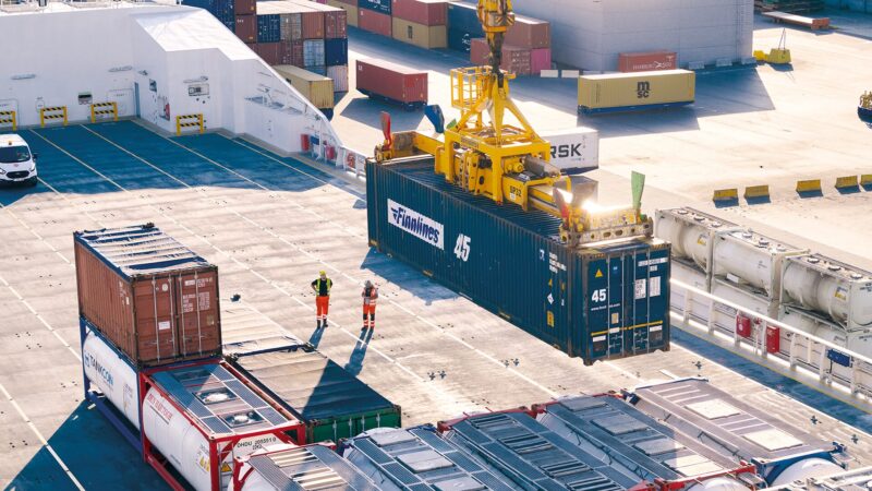 Overhead view of a busy shipping deck with a yellow crane lifting a dark blue container labeled "45" while two workers in high-visibility vests stand nearby among stacked containers.