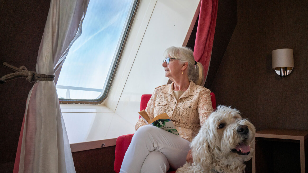 Dam med sin hund i en hytt ombord. Damen läser en bok och tittar ut genom fönstret.